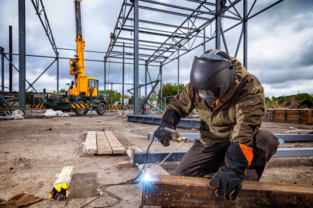 A young  man welder in brown uniform, welding mask and welders leathers, weld  metal  with a arc welding machine at the construction site, blue sparks fly to the sides